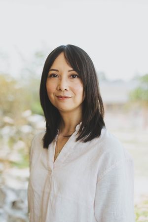 A woman with dark shoulder-length hair wearing a white linen top poses outdoors with a soft natural background.