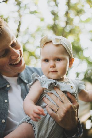 Smiling parent holding a cute baby girl in a blue ruffle outfit with a bow headband outdoors.