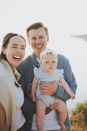 Happy family portrait of smiling parents holding their baby girl outdoors with a bright sky background.
