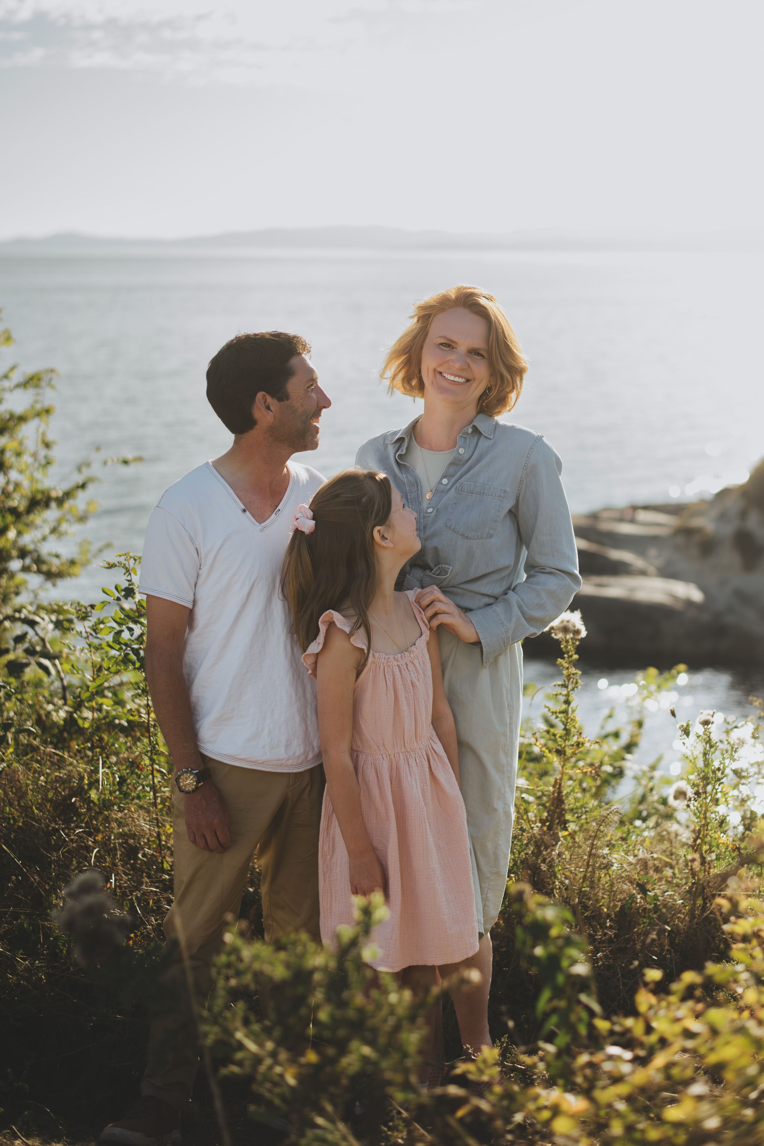 A happy family of three smiles together on a rocky coastal cliff at golden hour, with ocean views behind them.
