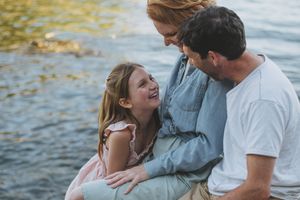 A happy family of three laughing together by a lake, with a young girl, red-haired mother, and father sharing a joyful moment.