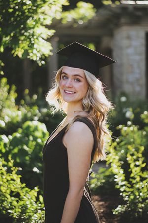 Smiling blonde woman in black graduation cap and gown posing outdoors among lush green foliage.