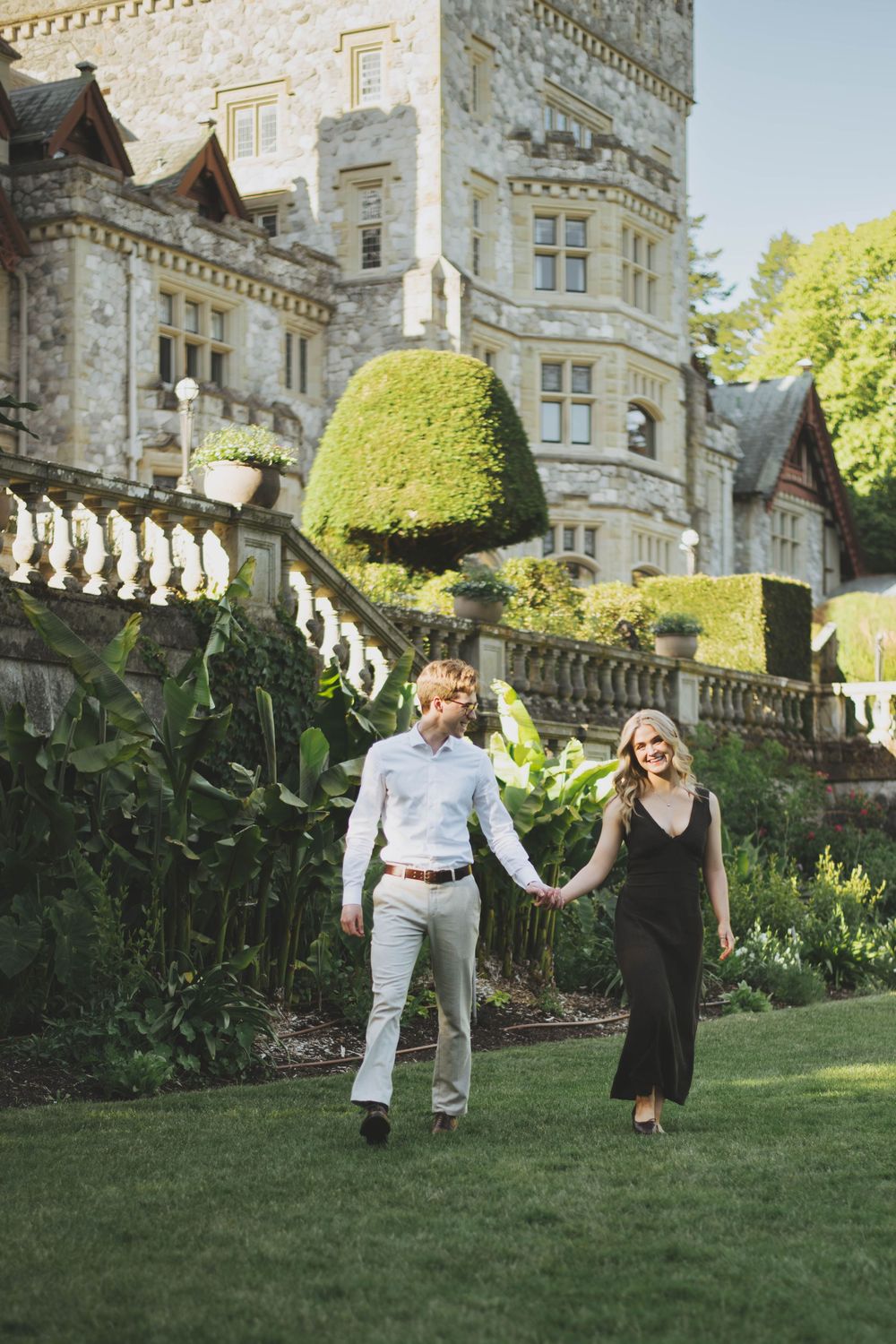 Couple holding hands walking on green lawn in front of a grand stone castle with manicured gardens.