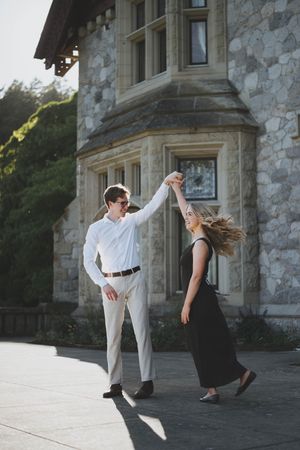 A couple dances outdoors in front of a stone mansion, man in white shirt spinning woman in black dress.
