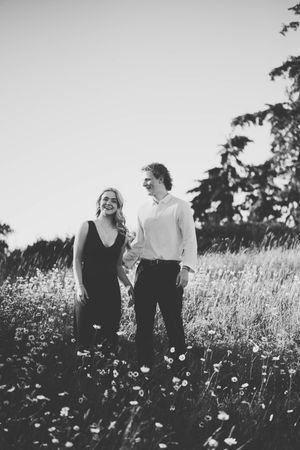 Black and white photo of a smiling couple holding hands in a wildflower meadow with trees in the background.