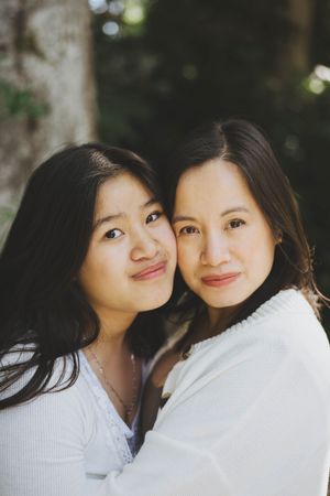 Two young Asian women with long dark hair smile warmly, cheeks together, wearing white tops outdoors.