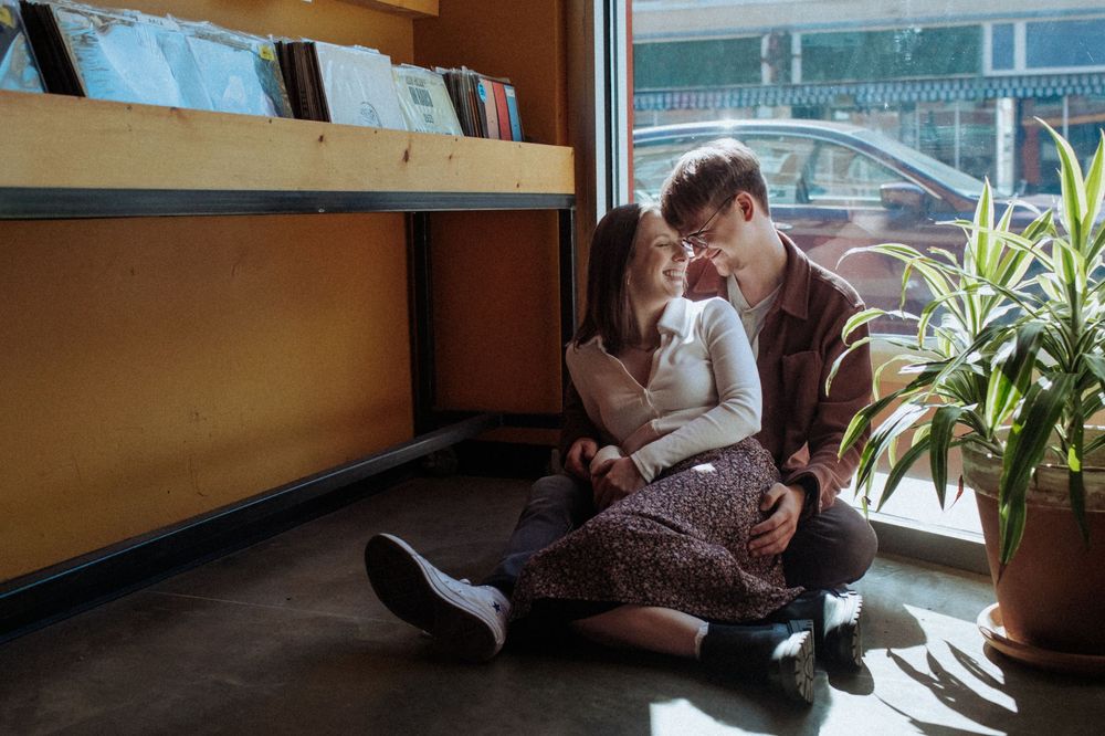Young couple in casual attire sitting intimately by window with potted plant in modern interior setting.