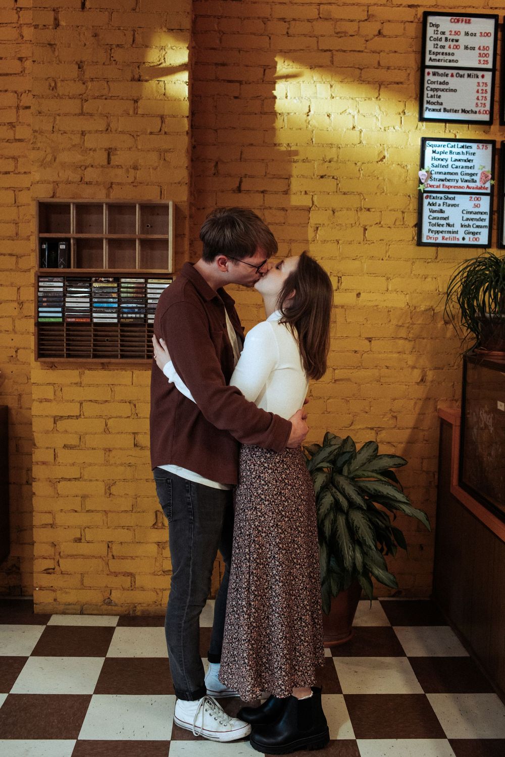 A couple embraces in front of a yellow brick wall with checkerboard flooring.