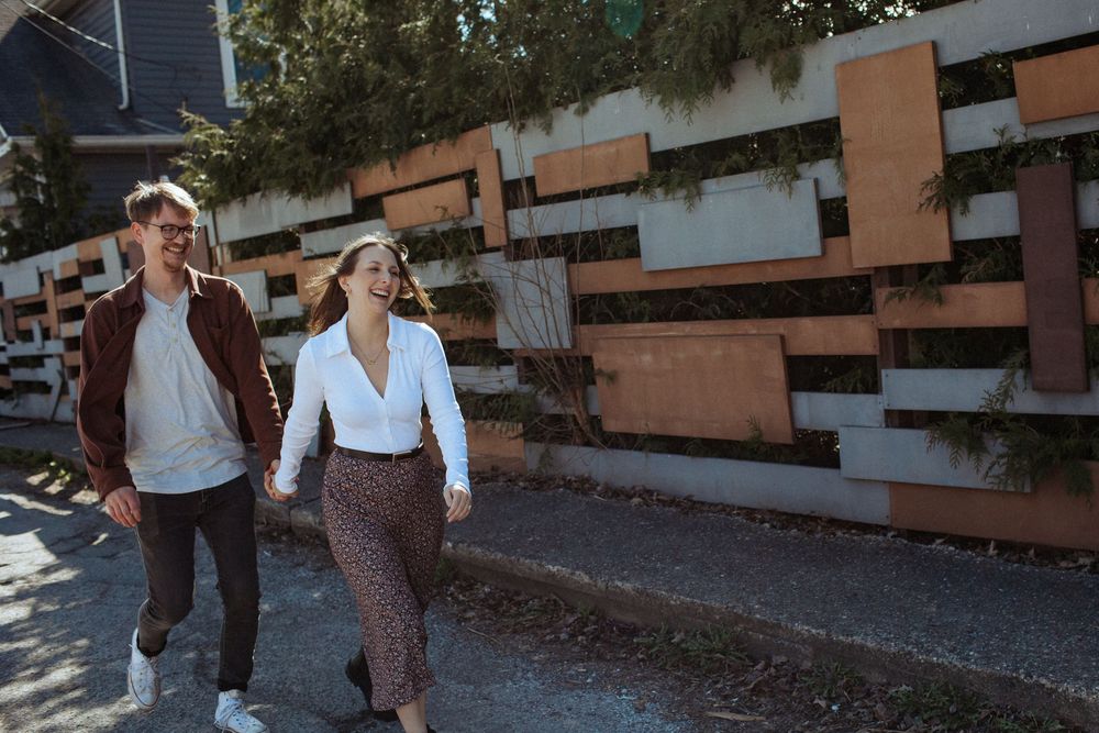 A couple walks playfully along a modern architectural wall with geometric patterns.