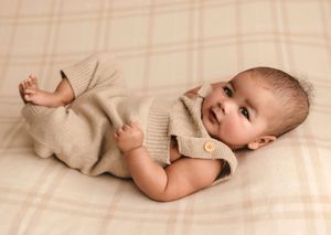 Baby boy in neutral knit overalls smiles as he lies against a soft plaid blanket, captured by Charlottesville baby and family photographer.