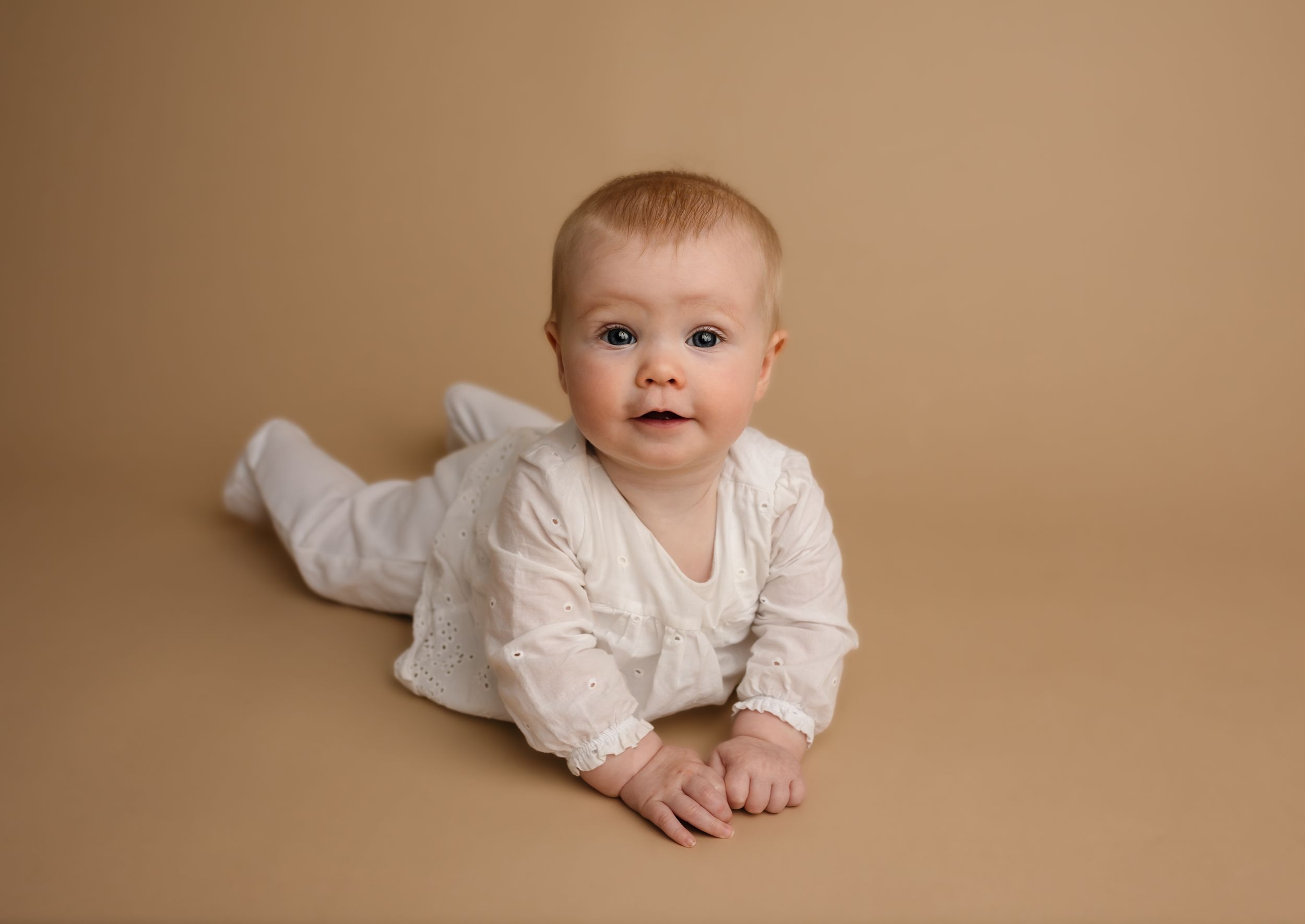 Smiling baby girl in white props up during tummy time on a warm neutral backdrop, captured by Charlottesville family photographer.