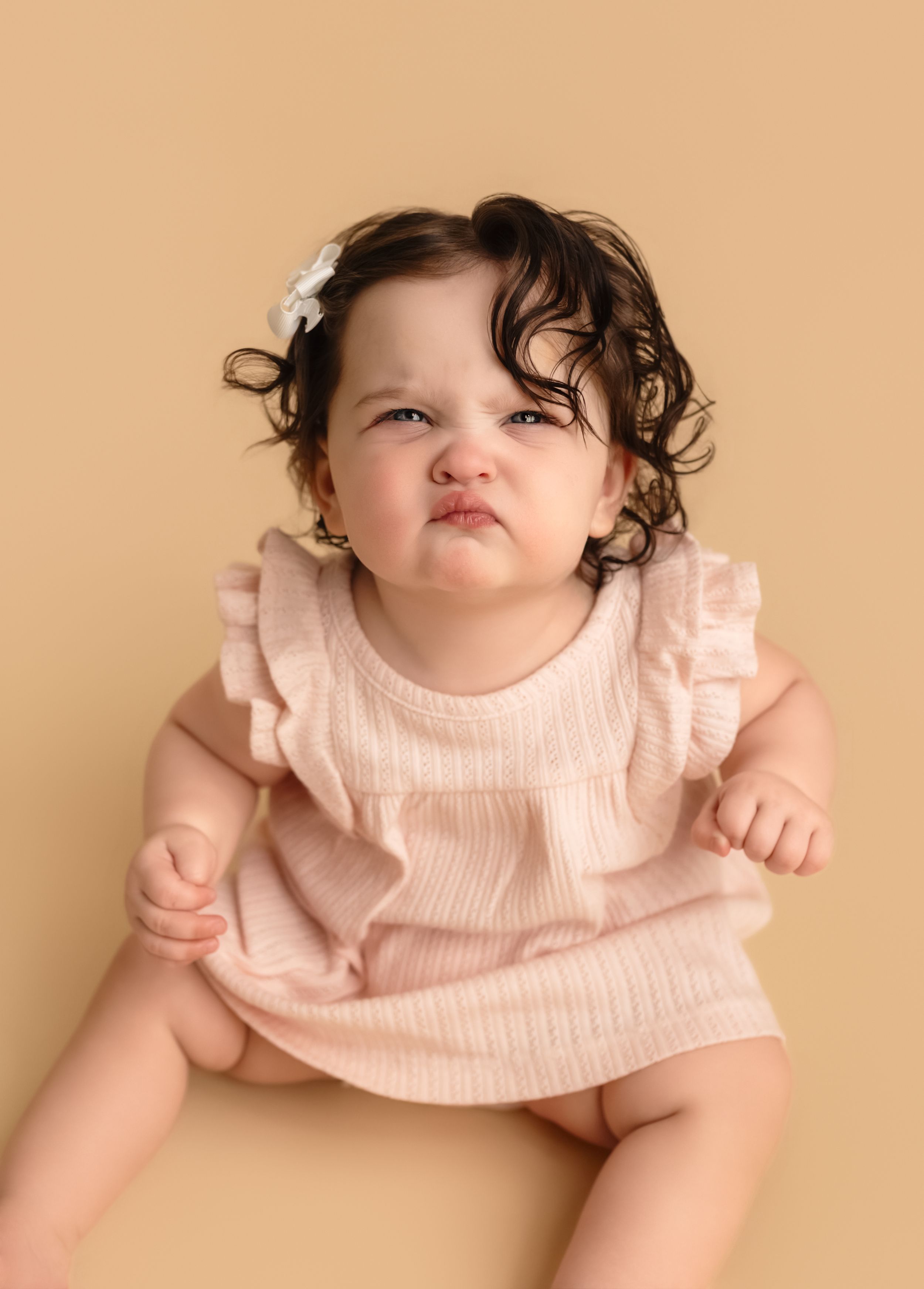 Curly headed baby girl makes faces at the camera as she sits against a warm, neutral backdrop at Charlottesville newborn and family studio.