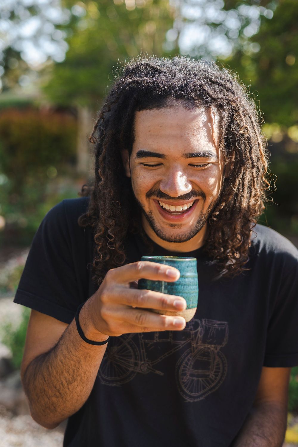 Person with curly hair wearing black t-shirt laughing while holding turquoise coffee mug outdoors.