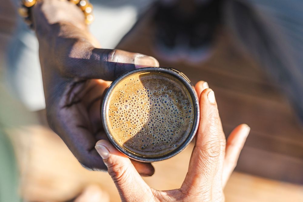 Hands holding a disposable coffee cup with frothy cappuccino against sunlit background.
