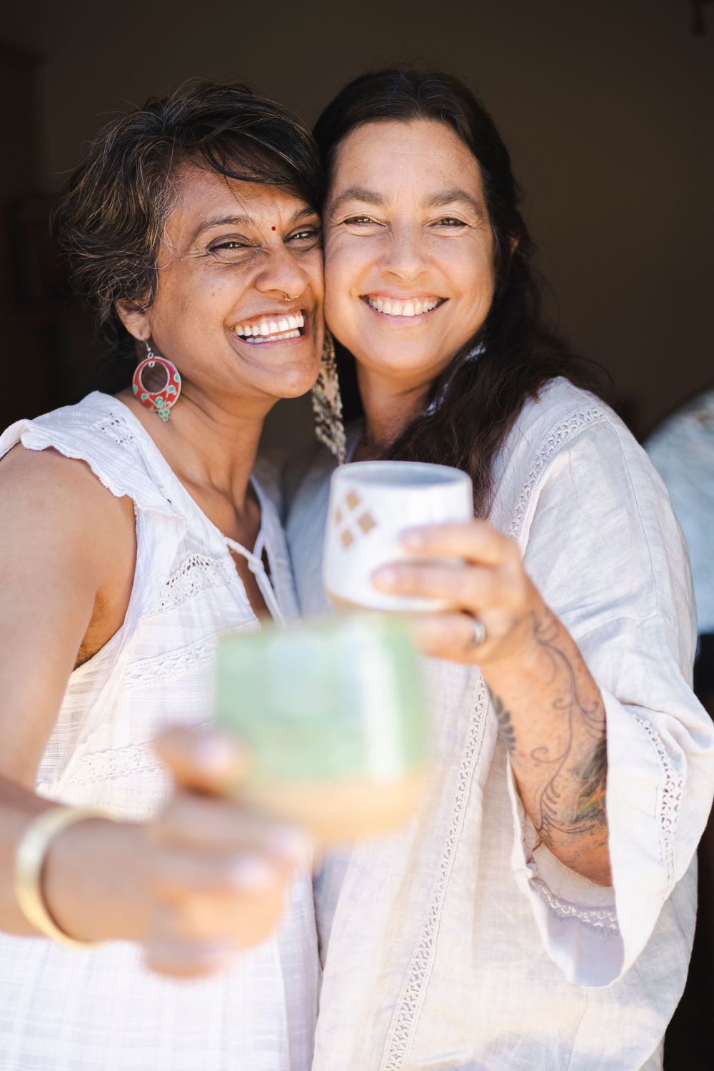 Two friends in white outfits share a joyful moment while toasting with colorful drinks at a casual gathering.