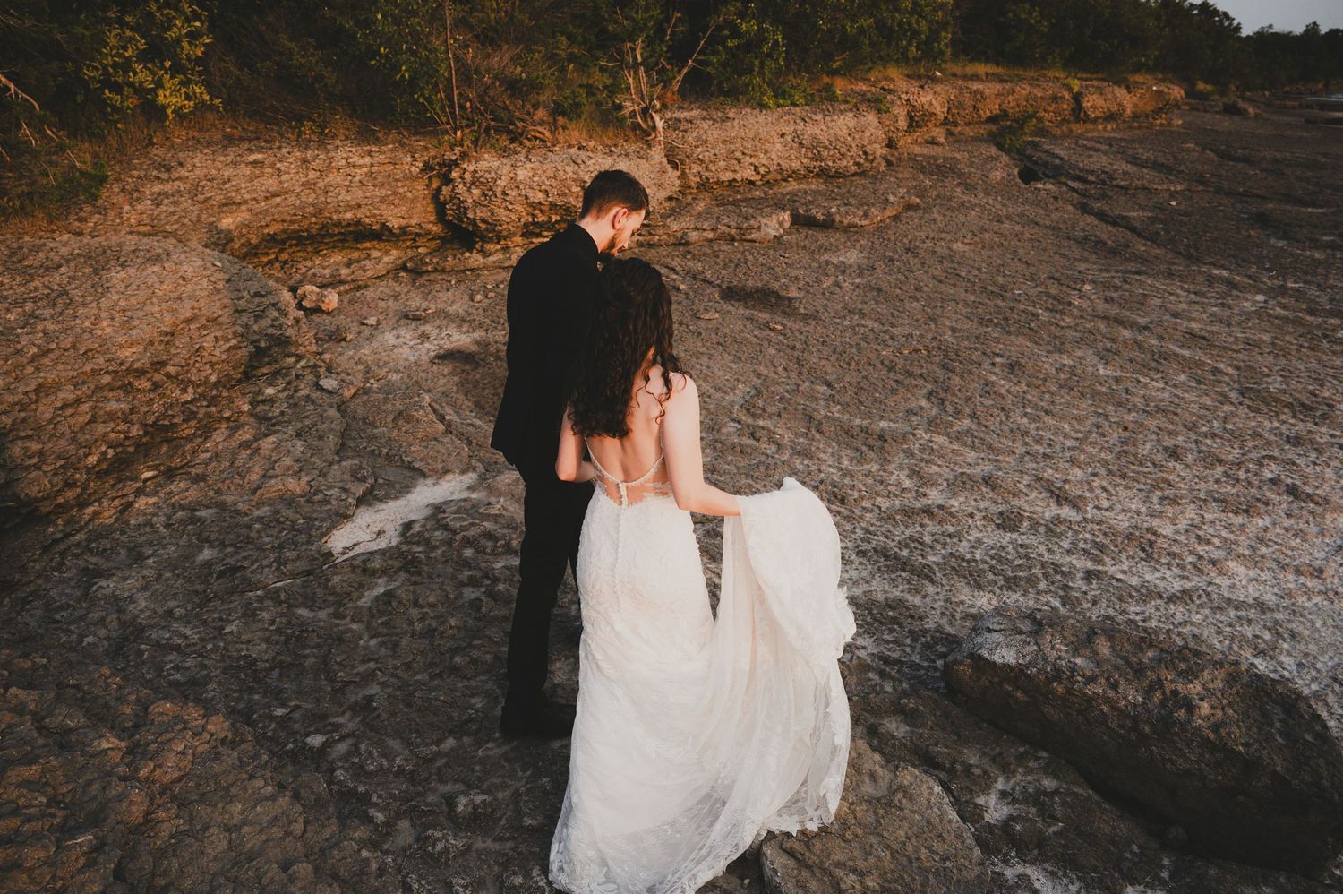 A couple in wedding attire shares an intimate moment on a rocky terrain during golden hour.