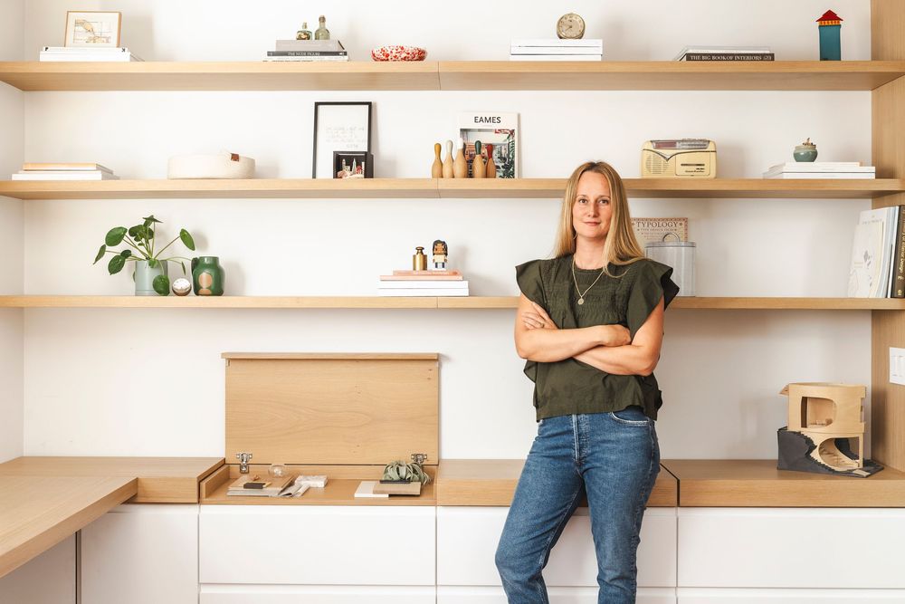 A person in jeans stands against a minimalist white wall with floating wooden shelves displaying decorative items.