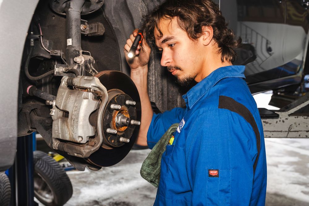 Auto mechanic in blue coveralls examines brake assembly and suspension components on a lifted vehicle.