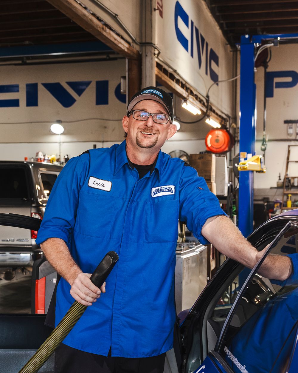 Auto mechanic in blue uniform smiling while working in a professional repair shop service bay.