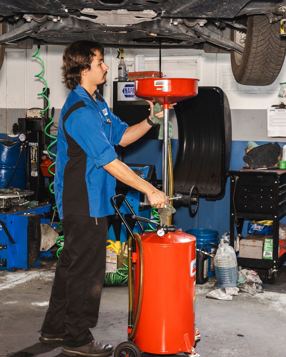 Auto mechanic in blue uniform uses a red oil drain container to perform an oil change service in a repair shop.