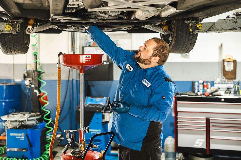 Auto mechanic in blue uniform performs maintenance inspection under vehicle lift in professional repair shop.