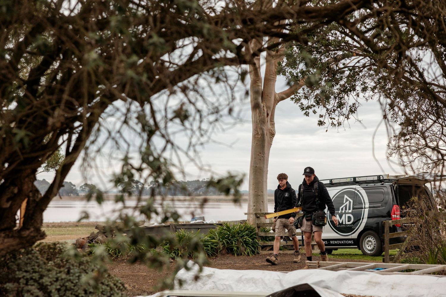 Two workers in black uniforms stand near a branded van outdoors by a waterfront, surrounded by trees and white tarps.