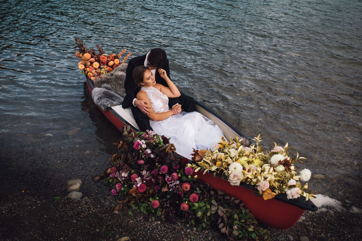 A romantic couple sits in a red canoe surrounded by floating flowers on dark rippling water during a photo session at Two Jack Lake in Banff