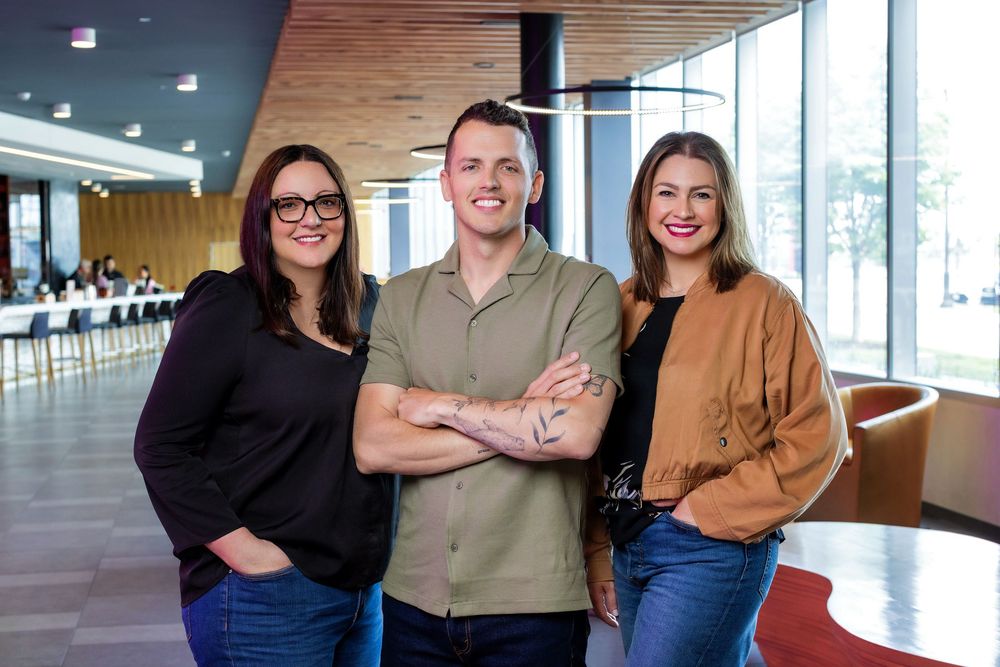 Smiling coworkers stand together in a modern office space with wooden ceiling and large windows.