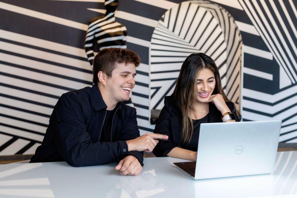 Two people laugh while looking at laptop together in modern office with black and white geometric wall pattern.