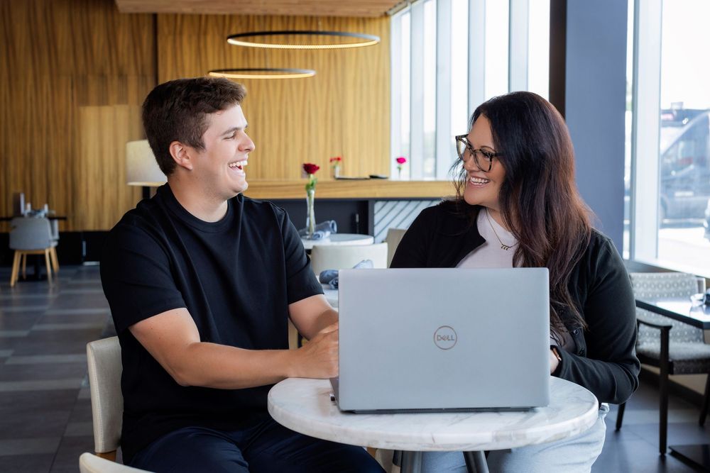 Two coworkers having a friendly discussion while working on a laptop in a modern office space with wooden walls.