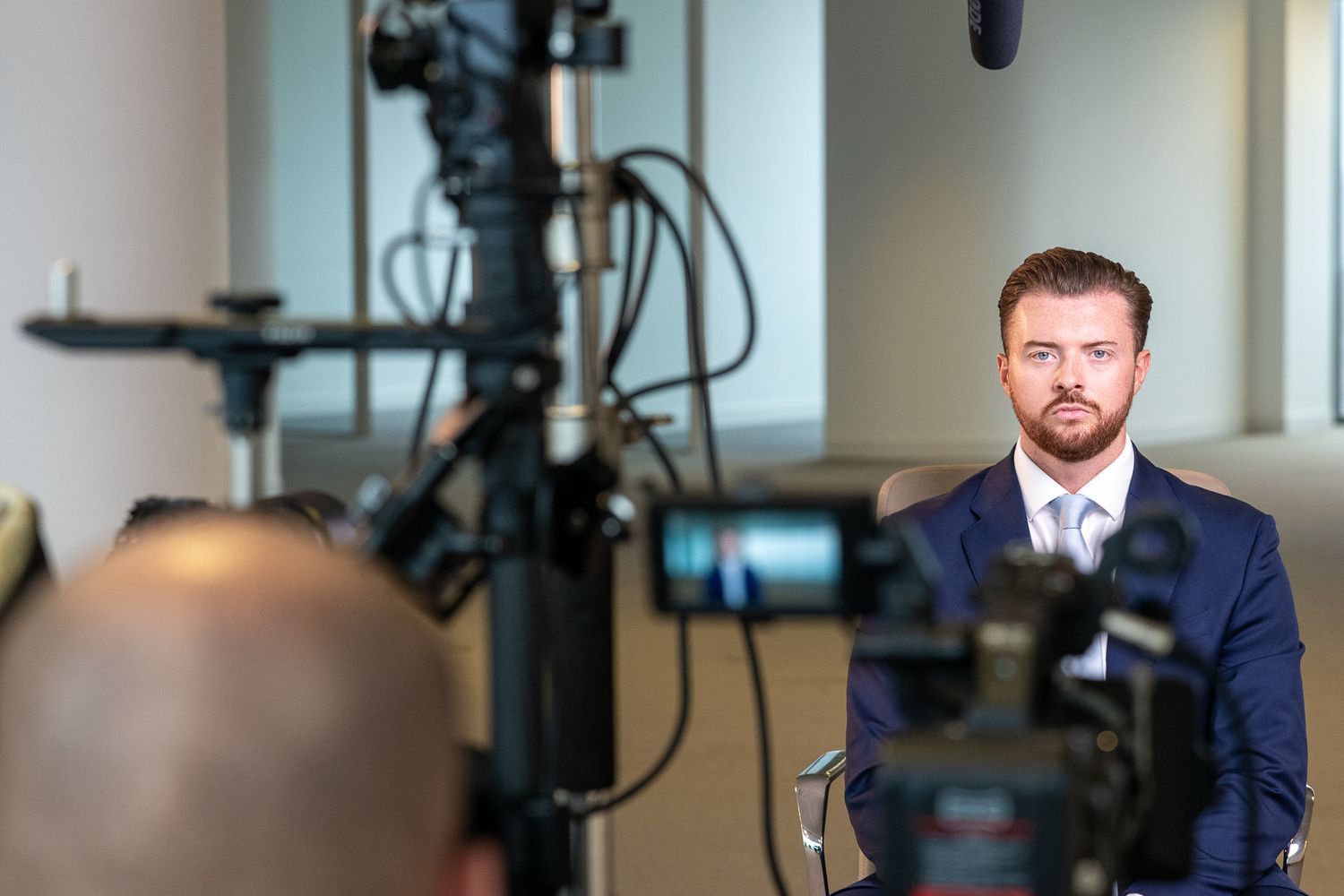 Professional wearing a navy suit speaks during an interview in a bright office setting with video production equipment.