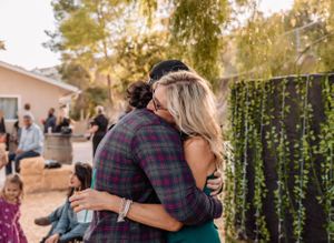 Guests embrace warmly at an outdoor party with string lights and trees in the background during golden hour.
