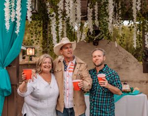 Friends raise their drinks in celebration under hanging floral decorations and teal drapery.
