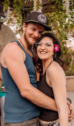 Romantic couple embraces outdoors under green foliage with turquoise curtains in the background.