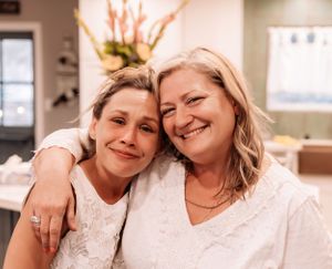 Two people in white outfits share warm embraces in an indoor setting.