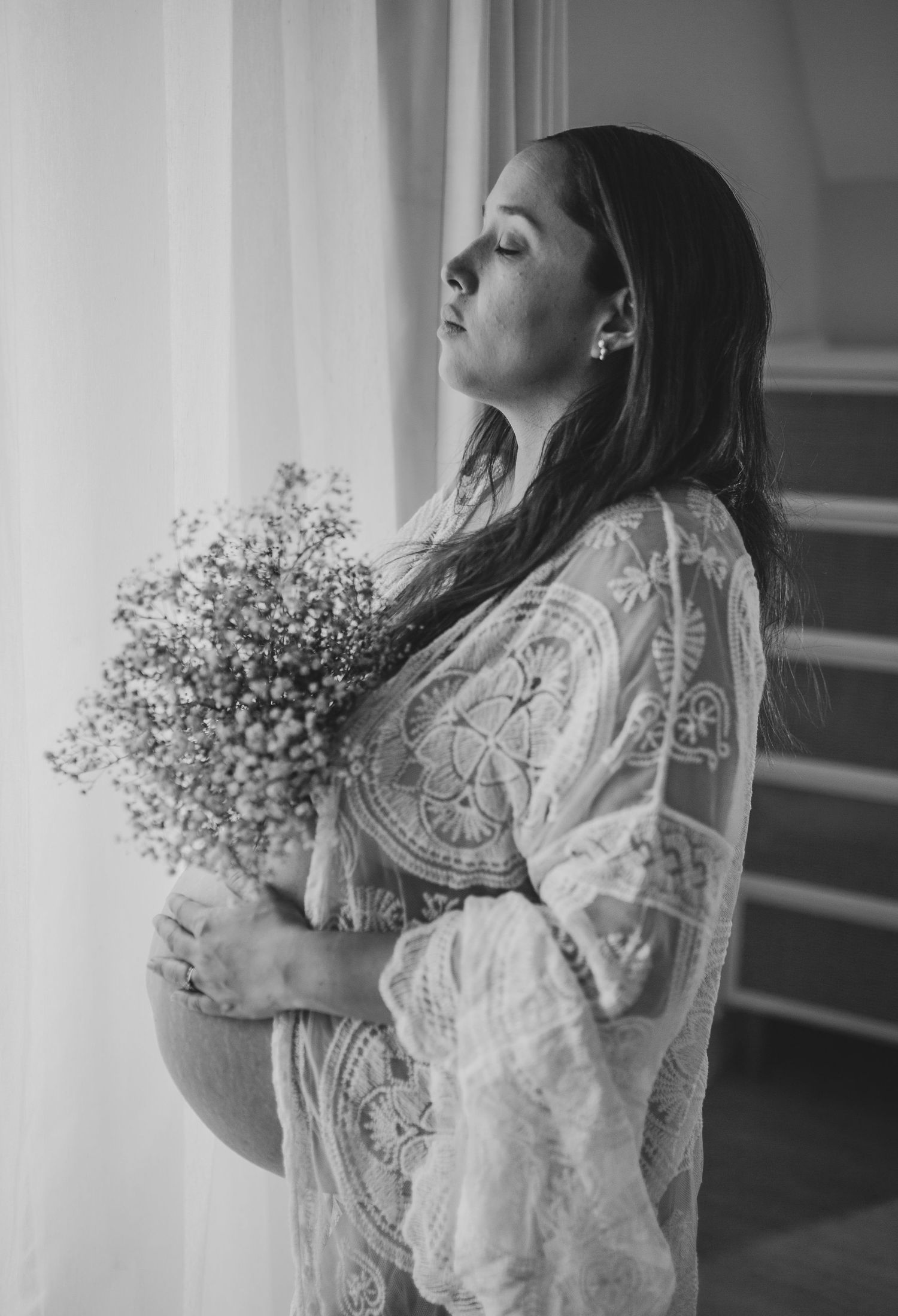 Black and white portrait of someone in a lace dress holding baby's breath flowers while standing by a window in soft light.