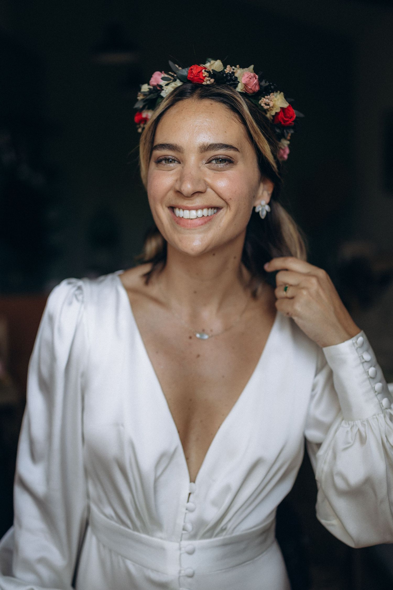 Beautiful bride wearing a white long-sleeve wedding dress and floral crown smiles warmly in a natural light portrait.