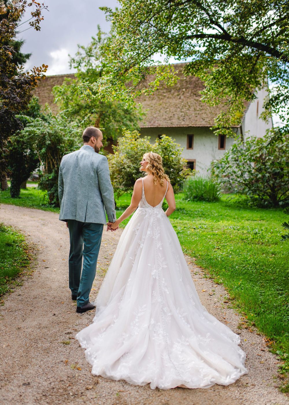 A couple walking hand in hand down a garden path with white dress and grey suit against lush green surroundings.