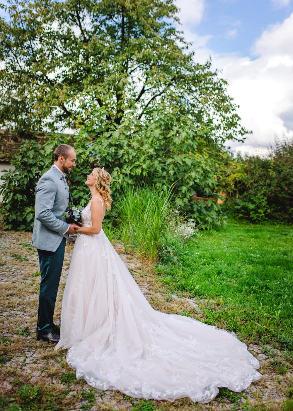 A romantic wedding couple stands together in a lush garden setting with a flowing white gown trailing behind them.