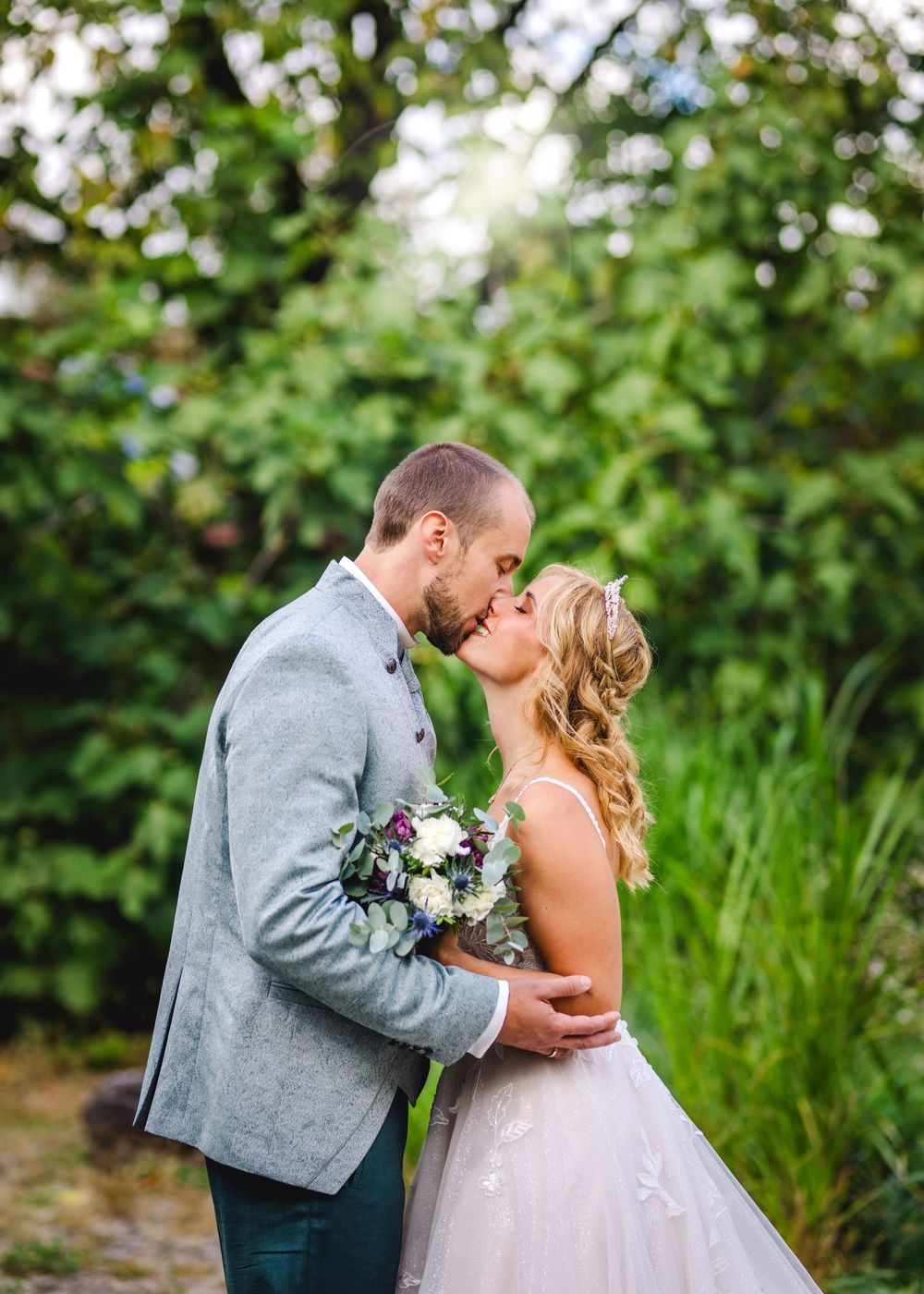 Newlyweds share a romantic kiss in a garden setting with lush greenery while holding a white and blue floral bouquet.