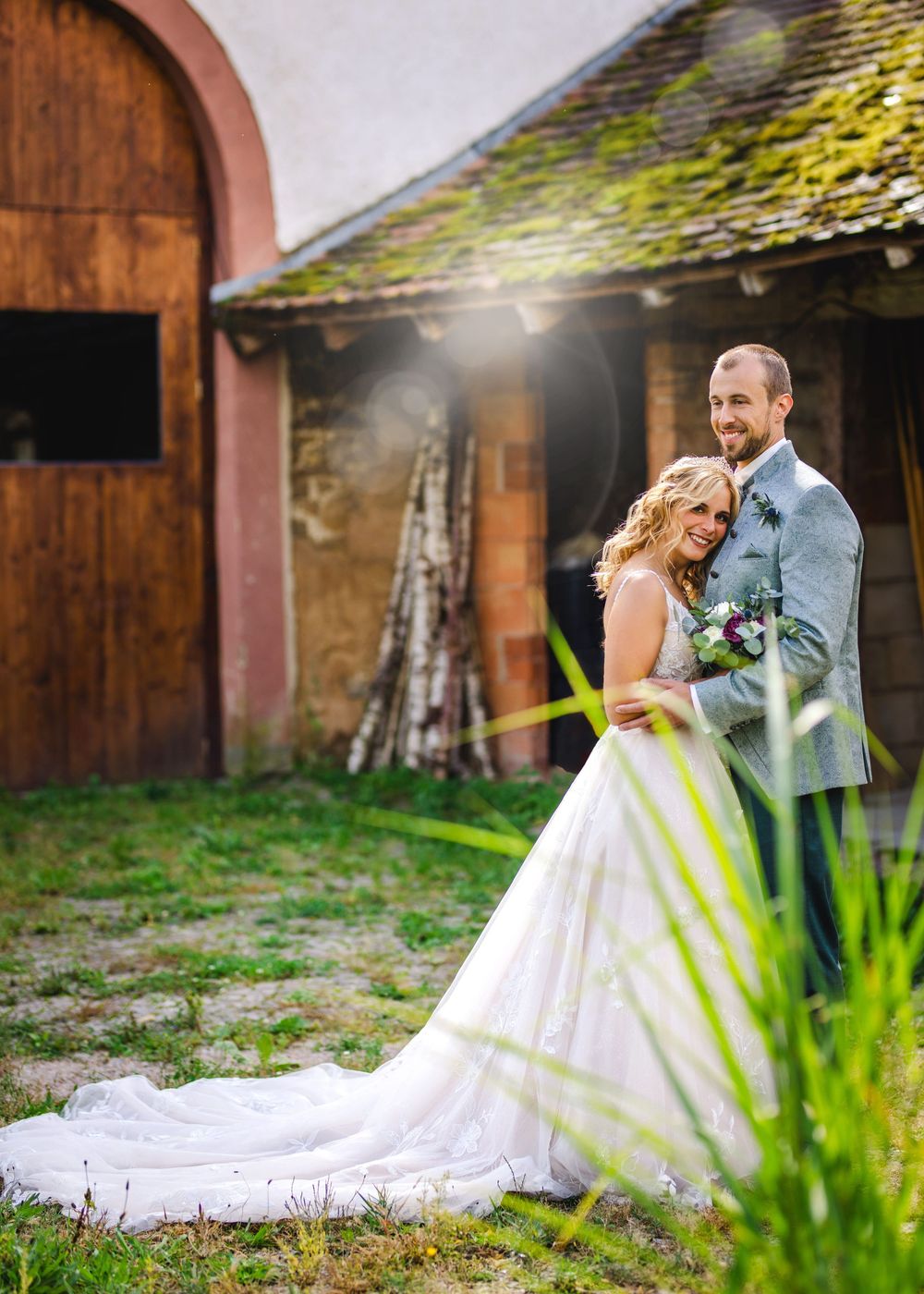 A couple shares a romantic embrace in their wedding attire against a rustic barn backdrop with sunlit moss-covered roof.