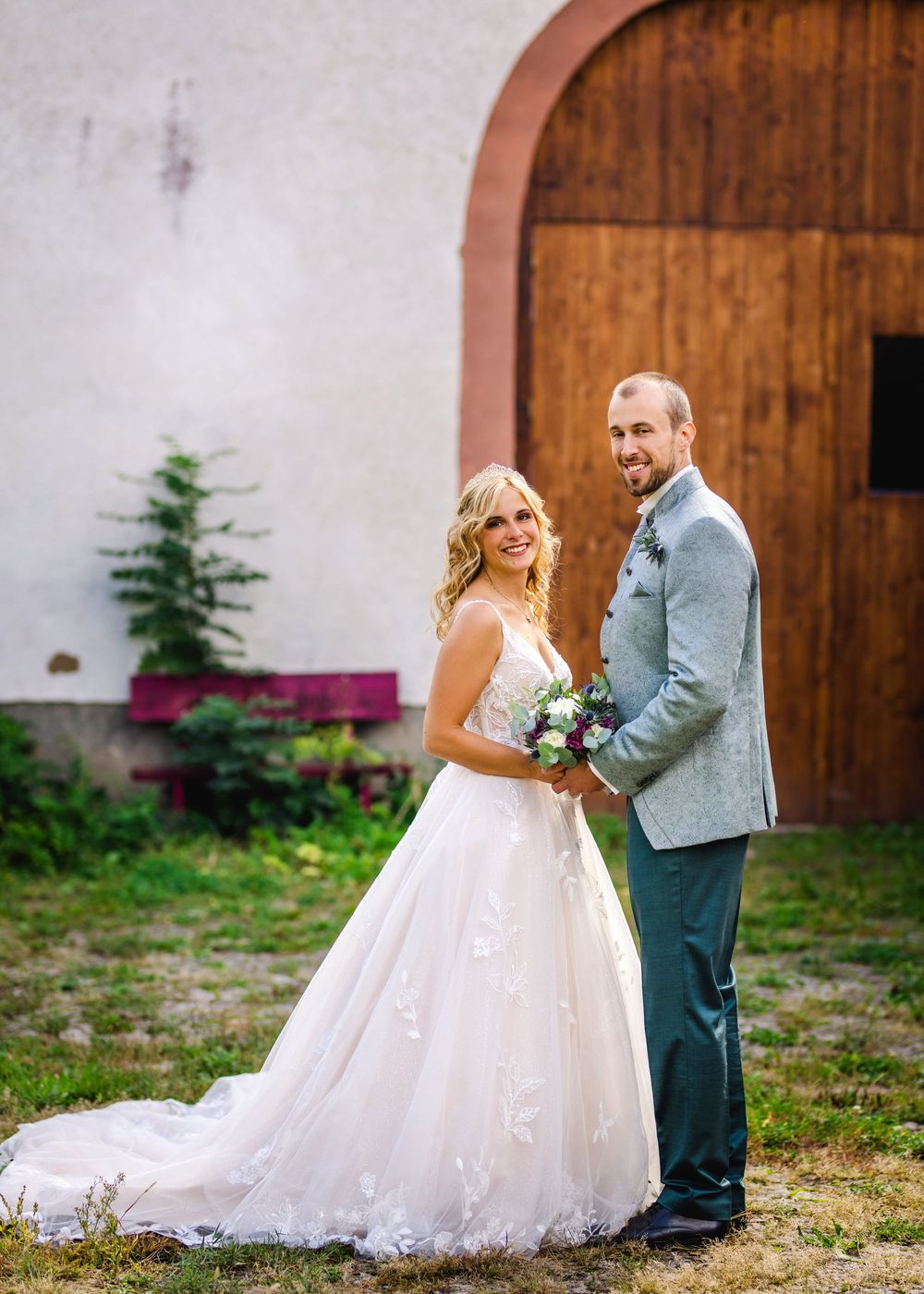 A romantic wedding couple poses together against a rustic wooden barn door in a garden setting.