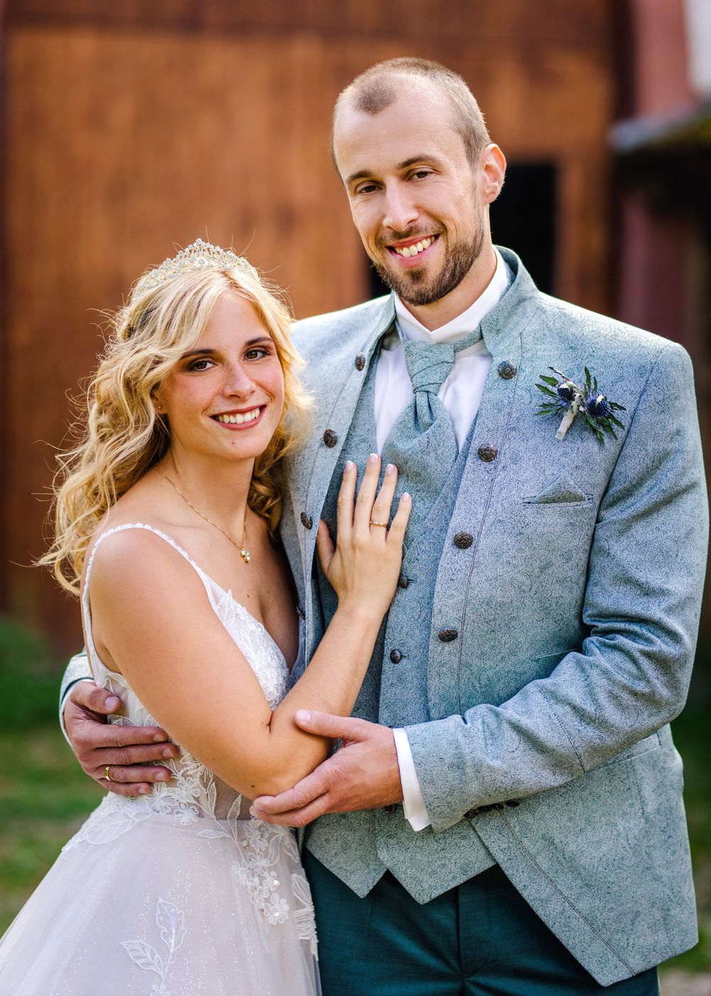 A bride and groom pose together in wedding attire, with the groom wearing a light gray suit and the bride in a sparkly gown.