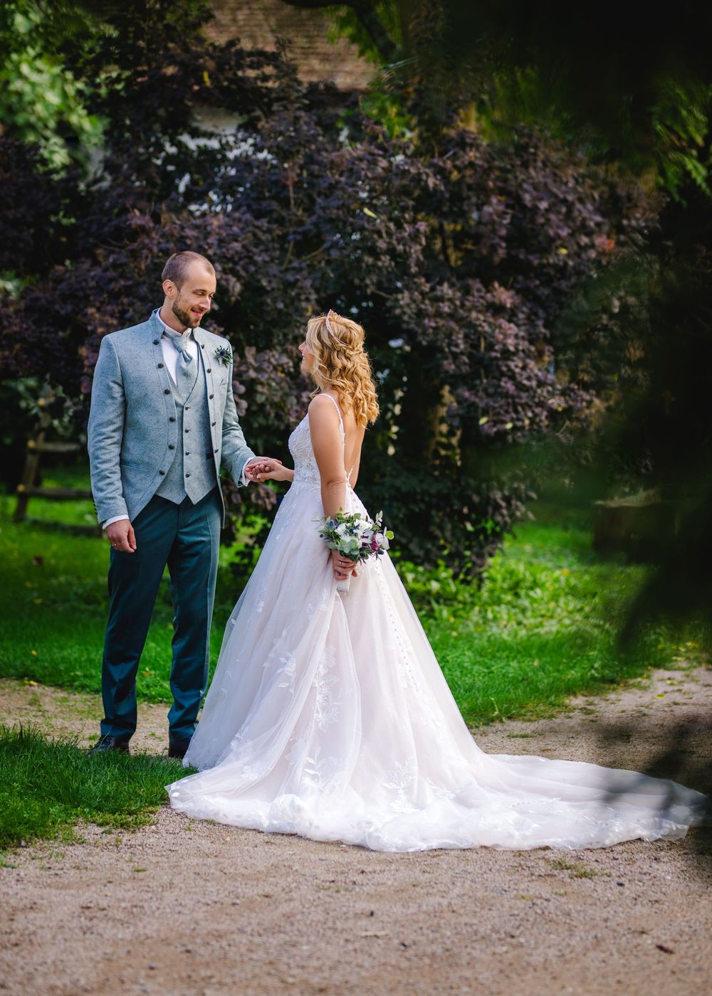 A couple in wedding attire walks together on a gravel path surrounded by lush green trees and foliage.