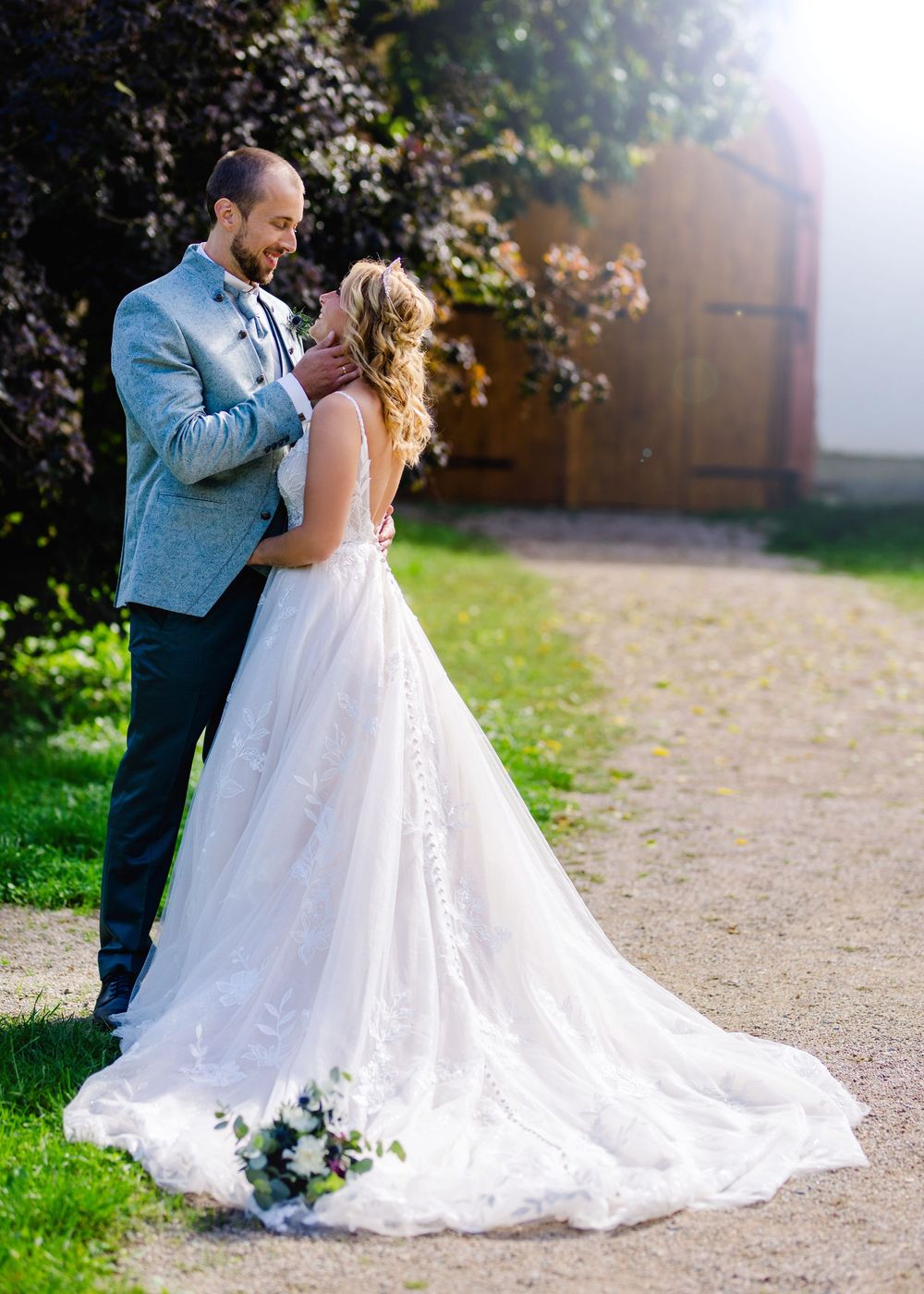 A romantic wedding couple embraces outdoors with a flowing white lace dress trailing behind them on a garden path.