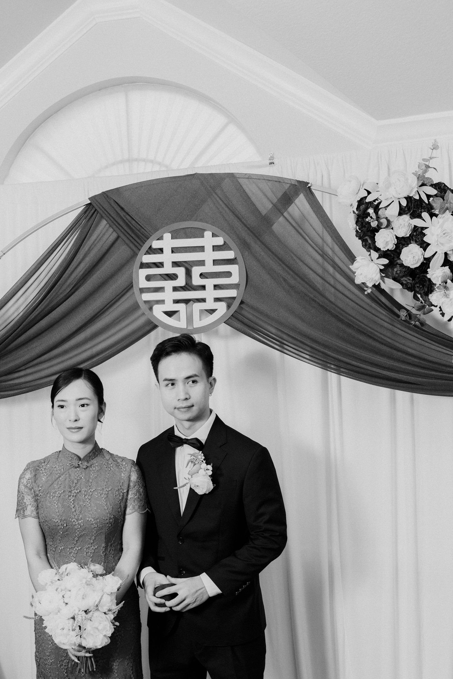 Couple poses beneath double happiness symbol at traditional Chinese wedding ceremony.