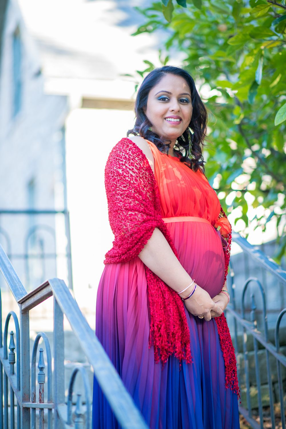 A person in a vibrant ombre dress with red lace shawl poses on outdoor steps near green foliage.