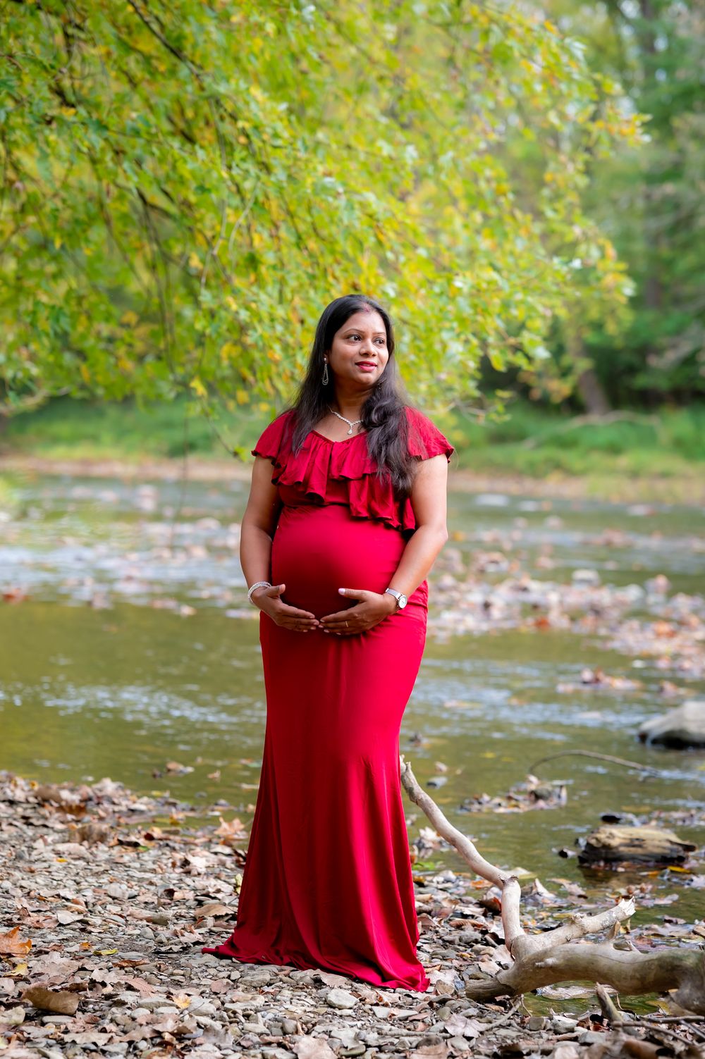 A person in a flowing red dress poses gracefully by a rocky creek surrounded by autumn foliage.