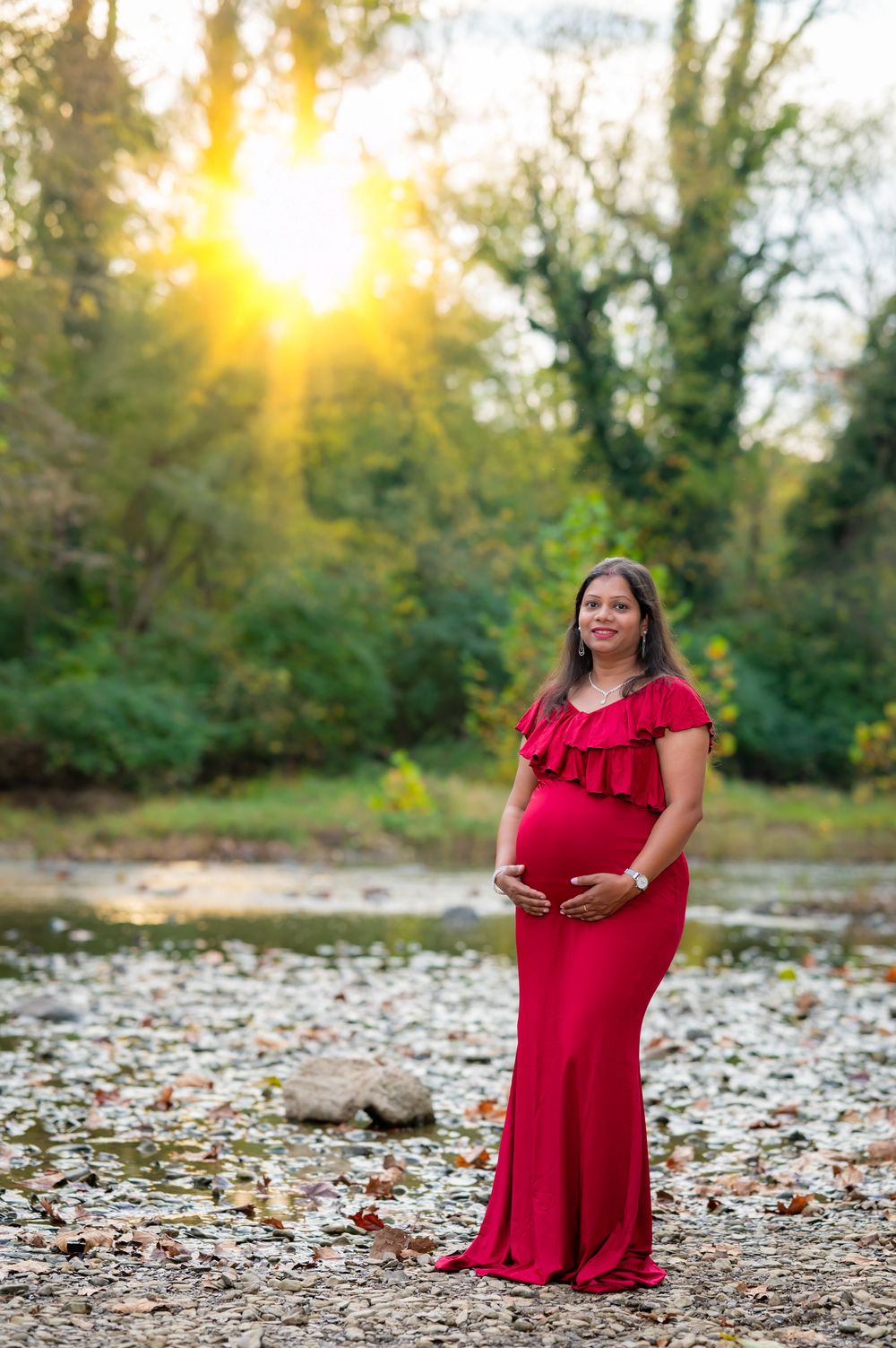 A maternity photoshoot at sunset by a river with autumn leaves scattered on the rocky shore.