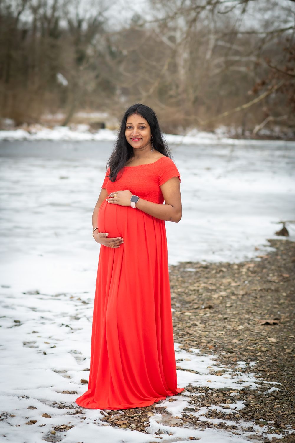 A person in a flowing red dress poses for maternity photos on a snowy beach during winter.